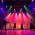 Scenă teatrală, lumini colorate, actori în rol.